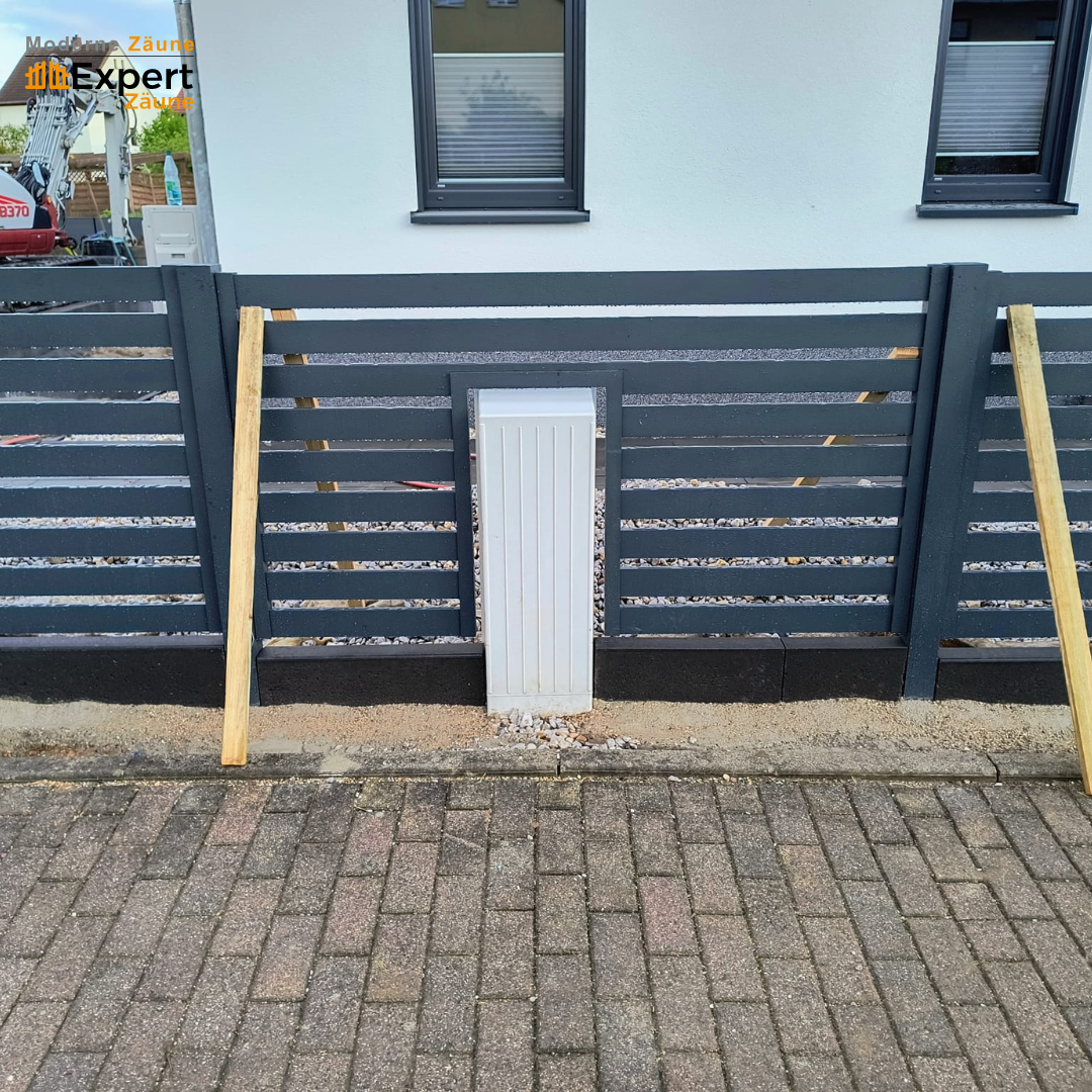 Nahansicht der präzisen Montage eines modernen Aluminiumzauns in mattem Anthrazit (RAL 7016). Ein passgenauer Ausschnitt im Zaunfeld integriert einen weißen Versorgungskasten perfekt in die Anlage aus 80x20 mm Profilen. Die auf Bordsteinen stehende Konstruktion wird durch Holzstützen stabilisiert und demonstriert die millimetergenaue Maßanfertigung von Moderne Zäune und Expert Zäune.